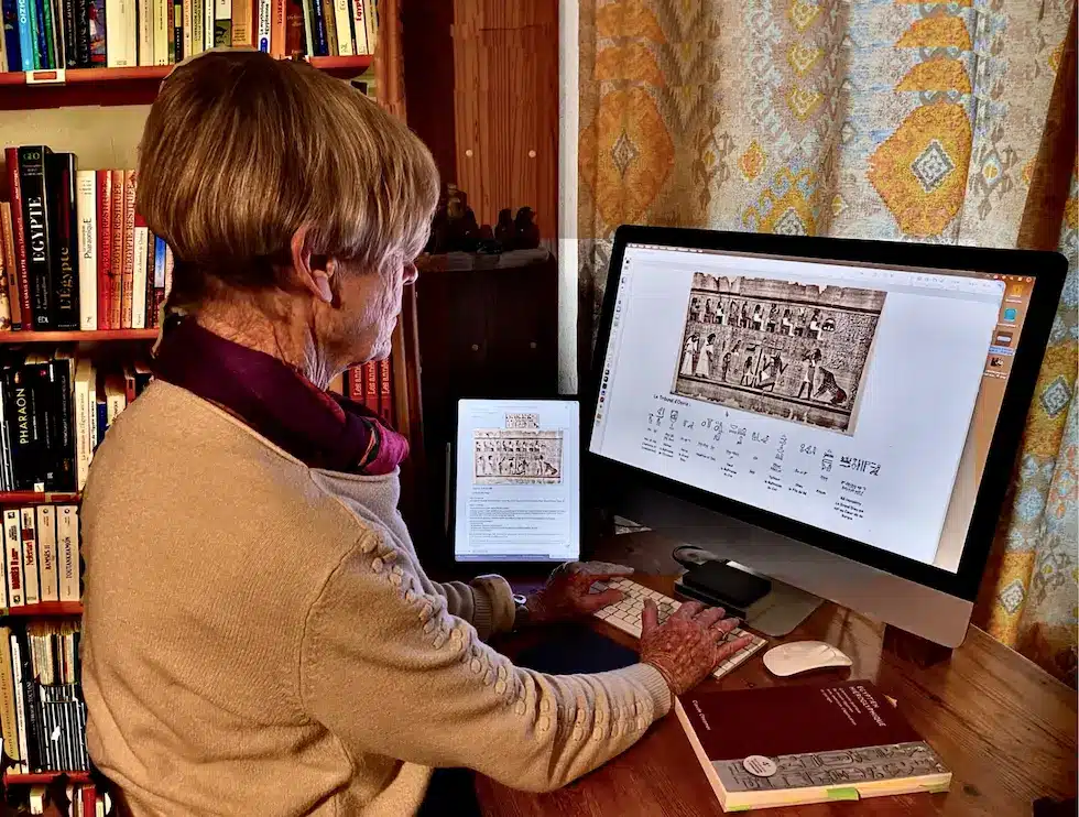 Photo d'une scribe moderne travaillant sur un ordinateur poss&eacute;dant un &eacute;cran grand format, avec, &agrave; ses c&ocirc;t&eacute;s, une tablette et un livre de grammaire d'&eacute;gyptien hi&eacute;roglyphique. Les &eacute;crans d'ordinateur et de tablette affichent l'image d'un papyrus et de sa traduction.