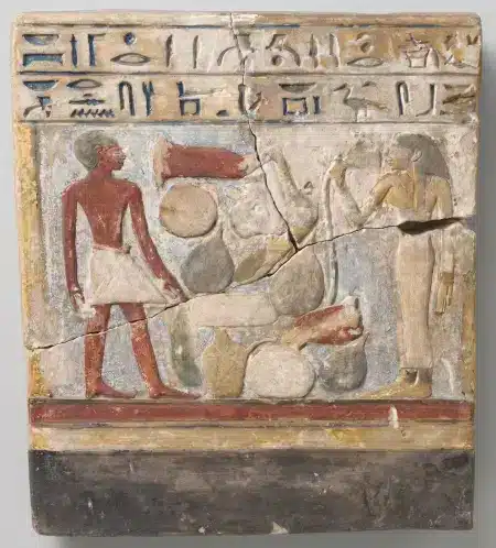 Photo de la stèle rectangulaire et polychrome représentant Sésostris et Satsobek debout de part et d’autre d’un amas d’offrandes. La scène est d’une part, surmontée de 2 lignes de hiéroglyphes gravés et peints en bleu, et d’autre part, soulignée dans la partie inférieure par des lignes épaisses de couleur : rouge, jaune puis une plus large encore, de couleur noire.